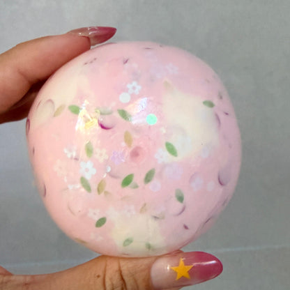 Handheld Sakura Cream Cheese squishy stress ball with thin skin, creamy sensory filling, pink floral details, and green leaf accents.
