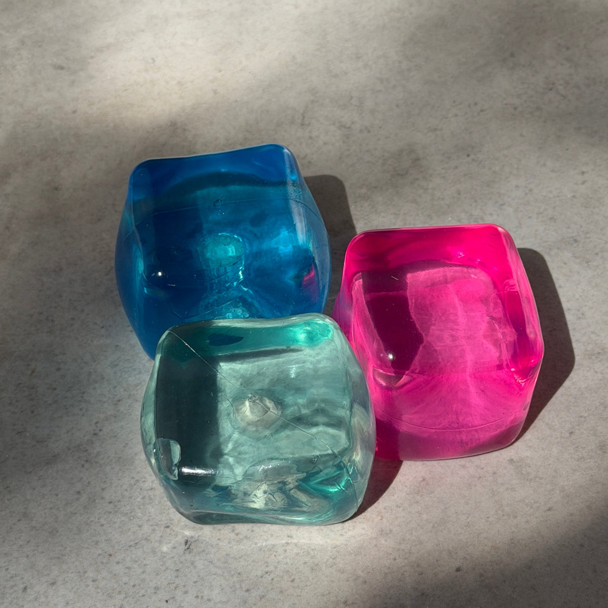 Three translucent cube squishy under natural lighting are blue, mint and pink color with square-shaped sensory and stress-relief toy designed to be squeezed, stretched, and kneaded before slowly returning to its original form